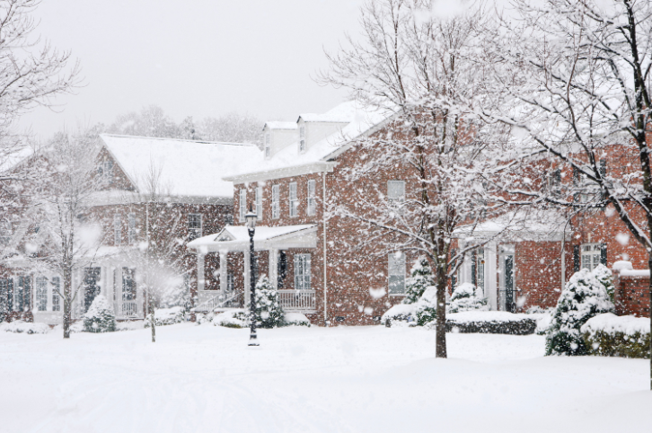 A row of brick and white houses in a residential neighborhood covered in freshly fallen snow, with trees and bushes dusted in white and snow continuing to fall.