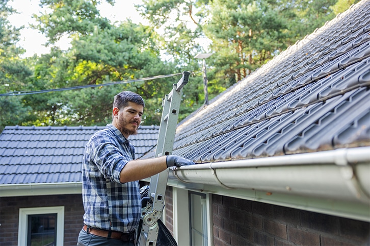 Man inspecting his roof and gutters by standing on top of a ladder.